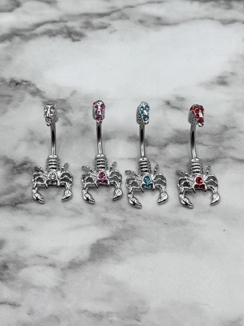 Pode incluir: Quatro an&eacute;is de umbigo de prata com pingentes de escorpi&atilde;o. Cada anel tem uma gema de cor diferente: rosa, azul, vermelho e transparente.