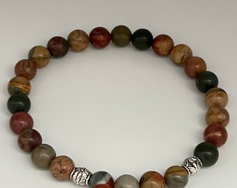 May include: A beaded bracelet featuring a variety of earth-toned jasper stones. The bracelet includes round beads in shades of brown, red, green, and gray, strung together with a stretch cord. Decorative silver-colored metal beads are interspersed throughout.
