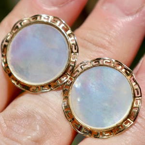 May include: A pair of round gold-toned cufflinks with a mother-of-pearl center. The cufflinks have a decorative openwork border. The iridescent centers reflect shades of blue, purple, and white. The cufflinks are shown against a blurred background.