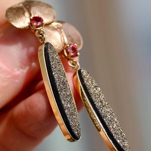 May include: Gold earrings with a black and gold textured oval pendant and a small red gemstone accent.