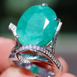 May include: A silver ring with a large, oval-shaped emerald gemstone surrounded by smaller diamonds. The ring has a unique, twisted design.