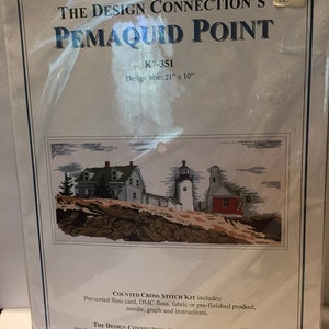 May include: A counted cross stitch kit by The Design Connection featuring a coastal scene with a lighthouse, houses, and a red building. The design size is 21" x 10". The kit includes pre-sorted floss, fabric, needle, graph, and instructions.