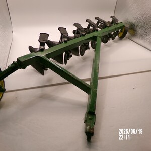 May include: A green metal agricultural plow toy featuring a triangular frame, multiple black plowshares, and yellow wheels. The device has a hitch point at the front and is angled to show the series of blades and the support structure clearly.