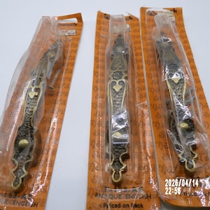 May include: Three antique brass-toned door pulls in their original packaging. The pulls have an ornate design with a textured surface. The packaging is orange and has the words "Antique English" printed on it.
