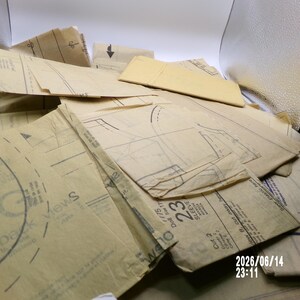 May include: A collection of tan sewing pattern tissue paper pieces featuring black printed lines, markings, and text including "vest back views" and "23 sleeve". The translucent paper sheets are layered and folded, showing various garment shapes.