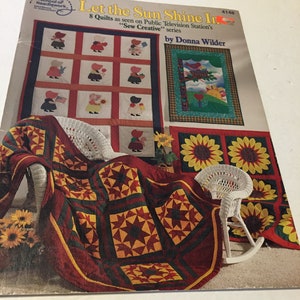 May include: A sewing pattern titled "Let the Sun Shine In" by Donna Wilder. The cover features images of quilts with various designs, including star patterns and scenes with figures. The pattern is from the American School of Needlework.