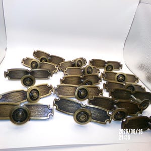 May include: A collection of antique brass-toned metal drawer pulls, each featuring an elongated rectangular backplate with a textured surface and a central circular knob. The hardware displays a dark, weathered finish with lighter highlights.