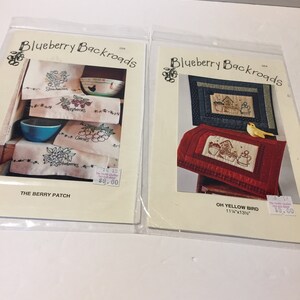 May include: Two sewing pattern packets titled Blueberry Backroads. The left packet, The Berry Patch, shows embroidered towels with fruit motifs. The right packet, Oh Yellow Bird, displays quilted wall hangings with birdhouse designs, measuring 11.25 by 13.5 inches.