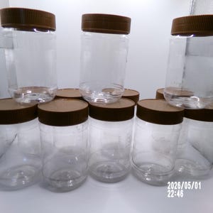 May include: A collection of clear plastic jars with brown screw-on lids. The jars are cylindrical and vary slightly in height. The lids are a solid, dark brown color. The jars are arranged in a group, some stacked on top of others.