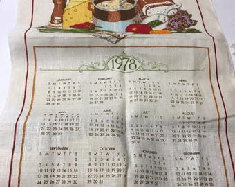 May include: A cream-colored fabric calendar towel for the year 1978. The top features a basket of vegetables, cheese, and a cooking pot with the text Bless This Kitchen with the joy of good food, the pleasure of welcome guests, and all the warmth of love.