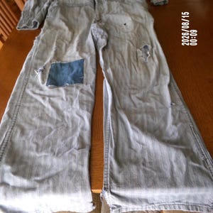 May include: A light gray long-sleeve jumpsuit featuring a button-up front, a collar, and long pant legs. The garment displays visible wear, including a dark blue denim patch on the right thigh and several small frayed holes on the legs.