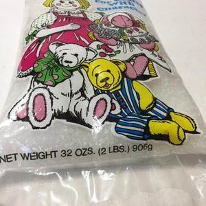 May include: A bag of white stuffing pellets for crafts with a doll, a teddy bear, and a bear wearing a blue and white striped shirt on the front. The bag says "For Crafts With Characters" and "Net Weight 32 ozs. (2 lbs.) 906g".