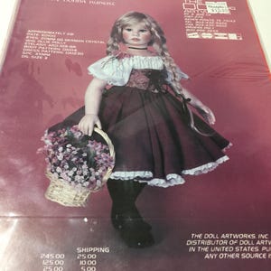 May include: A porcelain doll named Gretchen, wearing a burgundy dress with white lace trim and a white blouse with floral embroidery. She has blonde braids and is holding a wicker basket filled with pink flowers. The doll is approximately 20 inches tall.