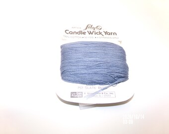 May include: A spool of Lily Candle Wick Yarn in slate blue color. The packaging is white with black text reading Lily Candle Wick Yarn, 100% cotton, 50 yds, 4 strands-3 ply, and 752 slate blue. The yarn is wrapped horizontally around the card.