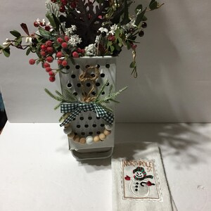 May include: A white metal container with a floral arrangement of red berries and greenery, adorned with a wooden bead garland and a green and white checkered bow. A white towel with a snowman embroidery that says "NORTHPOLE ST" is next to the container.