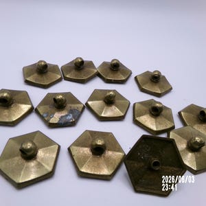 May include: A collection of twelve hexagonal brass buttons with a metallic gold finish. Each button features a central loop shank on the top surface. The buttons have a faceted geometric shape and show varying degrees of patina and surface wear.