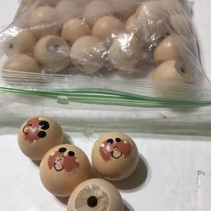 May include: A collection of round, light-colored wooden beads, some plain and some featuring hand-painted faces with black eyes, a small smile, and reddish-brown cheeks. The beads are contained within a clear plastic resealable storage bag.