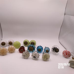 May include: A collection of decorative cabinet knobs in various shapes, colors, and materials, including clear glass, solid white, light green, blue, red, and beige. Each knob features a central metal mounting point with a circular base design.