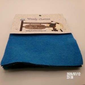 May include: A stack of bright blue felt fabric squares topped with a branded paper header card. The card features the text Wooly Charms, In The Patch DESIGNS, and WWW.INTHEPATCHWOOL.COM, along with illustrations of a small owl and a sheep.