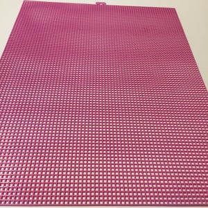 May include: A rectangular pink plastic canvas sheet featuring a uniform grid of small square holes throughout the entire surface. The sheet has a small, centered tab protruding from the top edge, designed for hanging or alignment purposes.