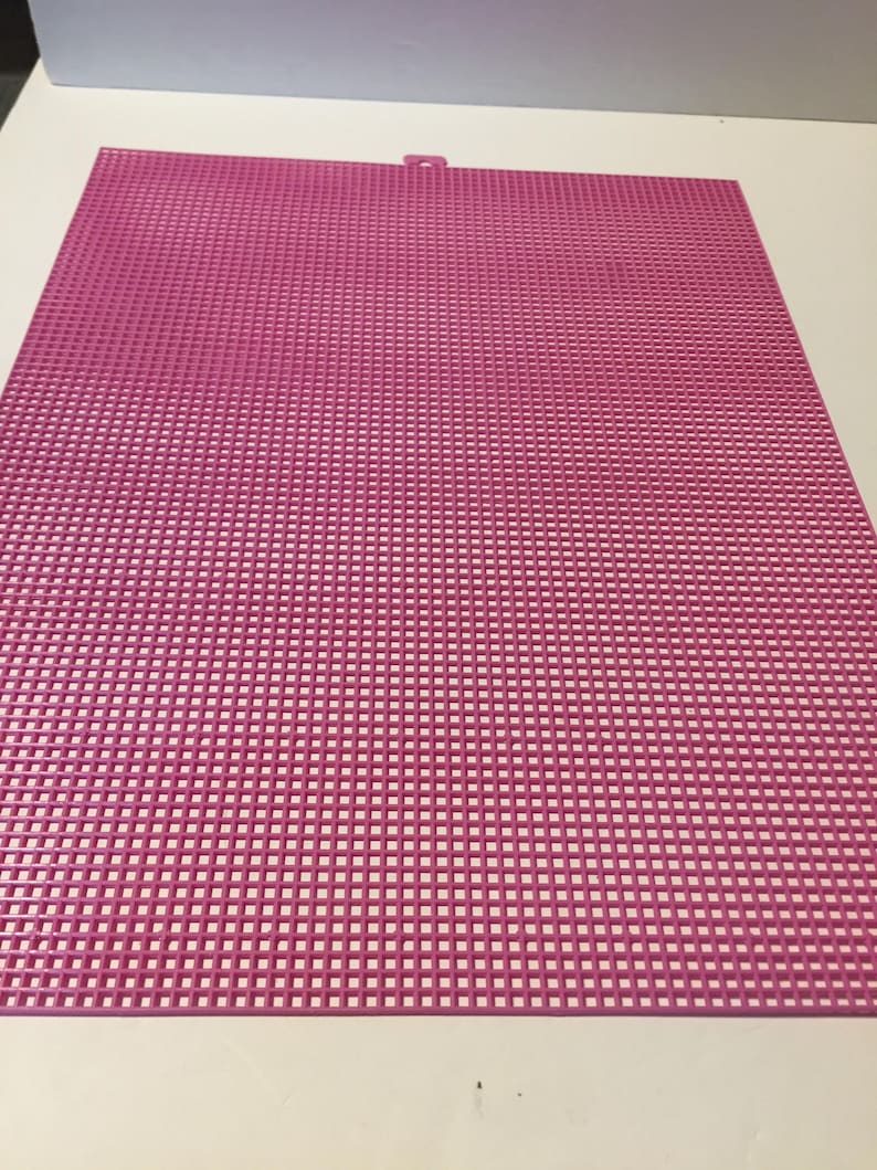 May include: A rectangular pink plastic canvas sheet featuring a uniform grid of small square holes throughout the entire surface. The sheet has a small, centered tab protruding from the top edge, designed for hanging or alignment purposes.