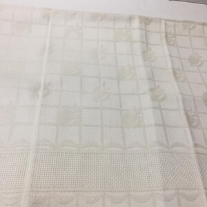 May include: A white kitchen towel featuring a woven grid pattern and a repeating jacquard design of small apples. The fabric includes a textured border with a scalloped edge detail along the bottom section, creating a subtle tonal effect.