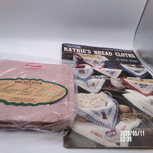 May include: A pink bread cover by Charles Craft, 18 x 18 inches, in a plastic package, next to a booklet titled "Kathie's Bread Cloths." The booklet features images of bread cloths with various designs and the date 2026/05/11.