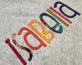May include: A gray fabric with the word "Sabrina" spelled out in colorful fabric letters. Each letter is outlined in a contrasting color.