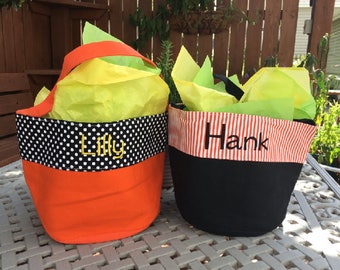 May include: Two fabric gift baskets. One is orange with a black and white polka dot band and the name "Lilly" embroidered in yellow. The other is black with a red and white striped band and the name "Hank" embroidered in black. Both are filled with yellow and green tissue paper.