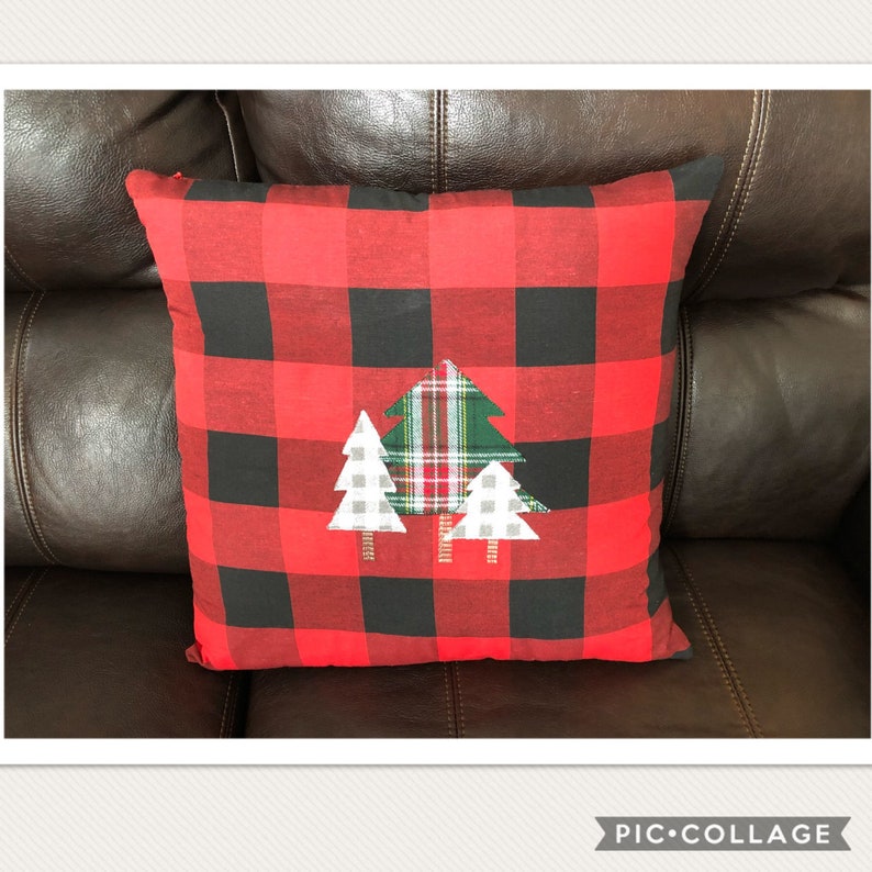 May include: A square decorative pillow with a red and black buffalo plaid pattern. Three appliqued Christmas trees in white, green, and plaid are centered on the pillow. The pillow is on a brown leather couch.