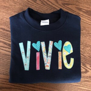 May include: A navy blue sweatshirt with the name "Vivie" spelled out in colorful fabric letters. Each letter is decorated with a different pattern and a small heart.