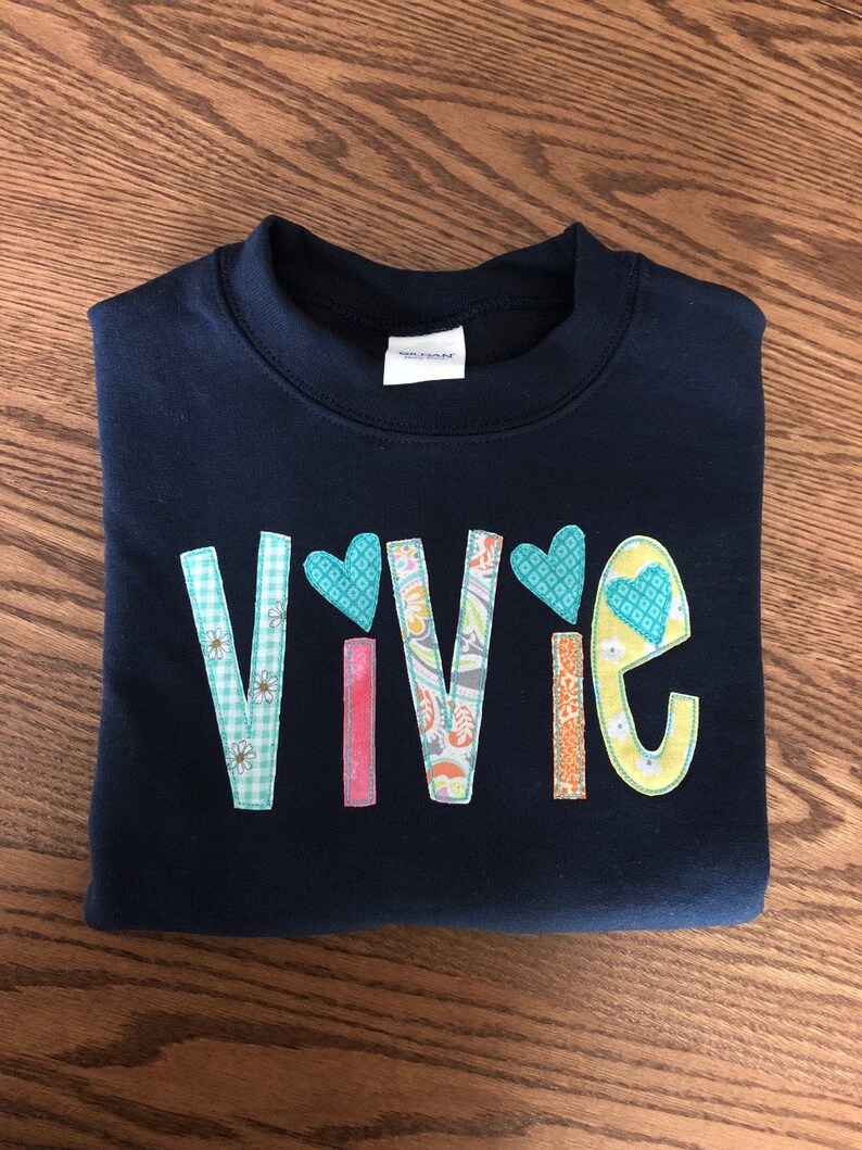 May include: A navy blue sweatshirt with the name "Vivie" spelled out in colorful fabric letters. Each letter is decorated with a different pattern and a small heart.