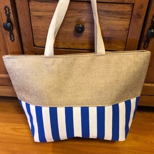 May include: A large, beige tote bag with blue and white striped fabric on the bottom half. The bag has two long, white straps.