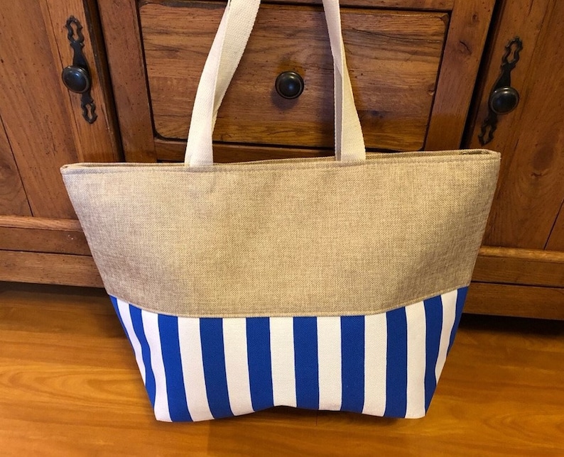 May include: A large, beige tote bag with blue and white striped fabric on the bottom half. The bag has two long, white straps.