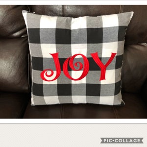 May include: Square pillow with a black, white, and gray buffalo check pattern. The word "JOY" is embroidered in red across the front. The pillow is resting on a brown leather couch.