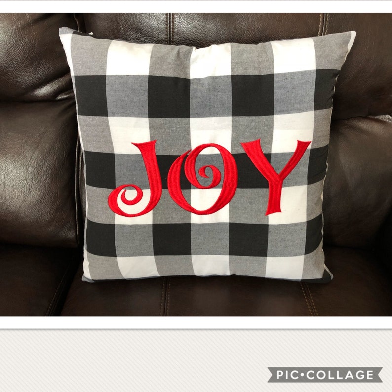 May include: Square pillow with a black, white, and gray buffalo check pattern. The word "JOY" is embroidered in red across the front. The pillow is resting on a brown leather couch.