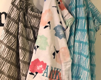 May include: Three towels hanging on a hook. The towel on the left is gray with white stripes. The towel in the middle is white with pink, blue, and green flowers and the word "ANK" embroidered on it. The towel on the right is turquoise with white stripes.
