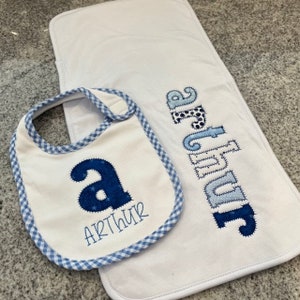 May include: A white baby bib with a blue and white checkered border and the name "Arthur" embroidered in blue. The bib has a large blue letter "A" in the center. A white burp cloth with the name "Arthur" embroidered in blue patchwork letters.