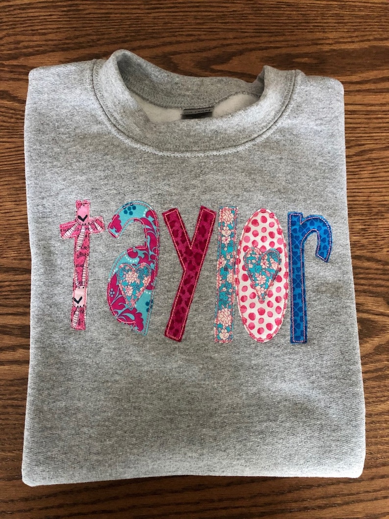 May include: Gray sweatshirt with the name "taylor" spelled out in colorful fabric letters. Each letter is made of different patterned fabric with pink, blue, and white accents.