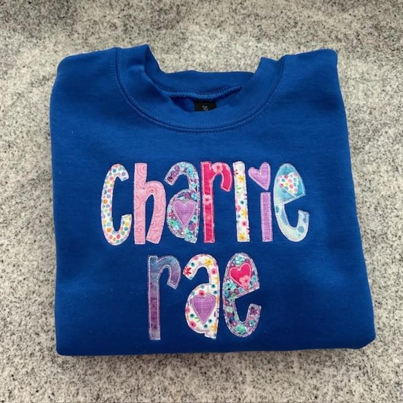 May include: A royal blue sweatshirt with the name "charlie rae" spelled out in colorful fabric letters with hearts and floral patterns.