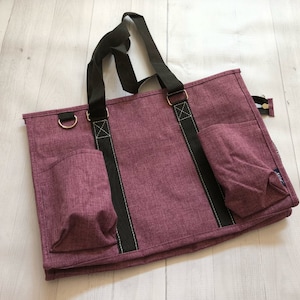 May include: A purple tote bag with black straps and two zippered pockets. The bag has a textured fabric and is made of a durable material.