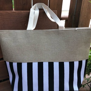May include: A large beige and black striped tote bag with white canvas handles. The bag is made of a woven fabric and has a zipper closure.