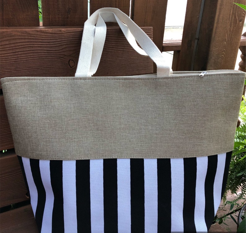 May include: A large beige and black striped tote bag with white canvas handles. The bag is made of a woven fabric and has a zipper closure.