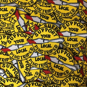 May include: A pile of yellow embroidered patches. Each patch displays the text "SUPPORT YOUR LOCAL PINSTRIPER" in black, with a knife design and red details. The patches are circular in shape and densely packed together.