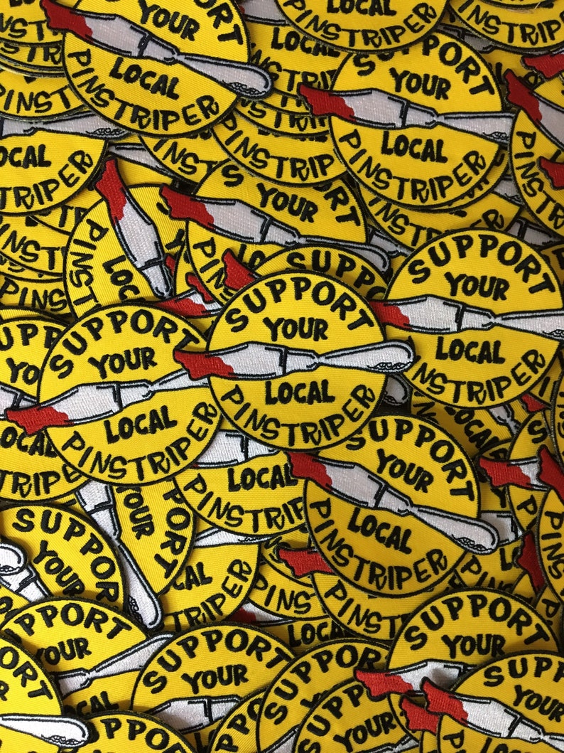 May include: A pile of yellow embroidered patches. Each patch displays the text "SUPPORT YOUR LOCAL PINSTRIPER" in black, with a knife design and red details. The patches are circular in shape and densely packed together.