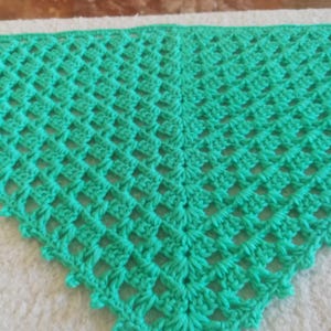 Op de afbeelding: Een handgemaakte, turquoise gehaakte sjaal met een ruitvormig patroon. De sjaal is gemaakt van zacht garen en heeft een geschulpte rand. De open weving zorgt voor een licht en luchtig gevoel.