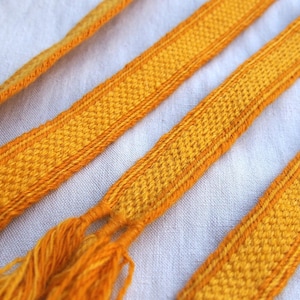 May include: Close-up of several woven yellow and orange ribbons with a textured pattern. The ribbons have a fringed end and are displayed on a white fabric background. The ribbons are likely used for decorative purposes.