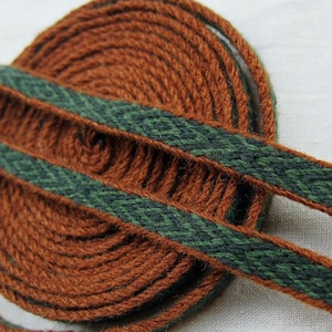 May include: A close-up of a spool of brown and green woven ribbon. The ribbon has a geometric pattern and is coiled neatly.