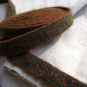 May include: A brown and green woven trim with a geometric pattern. The trim is rolled up and is on a white fabric background.
