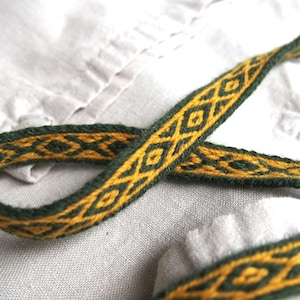 May include: A close-up of a green and gold woven ribbon with a geometric pattern. The ribbon is lying on a white fabric surface.